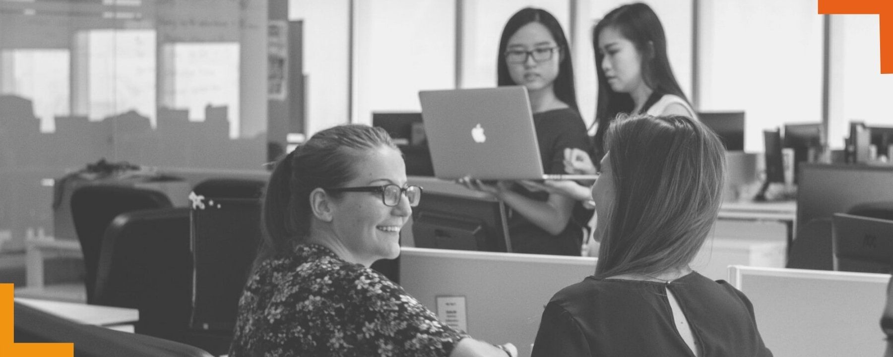 Two women in open office in foreground seen in profile chatting. Two other women in background focused on a laptop.