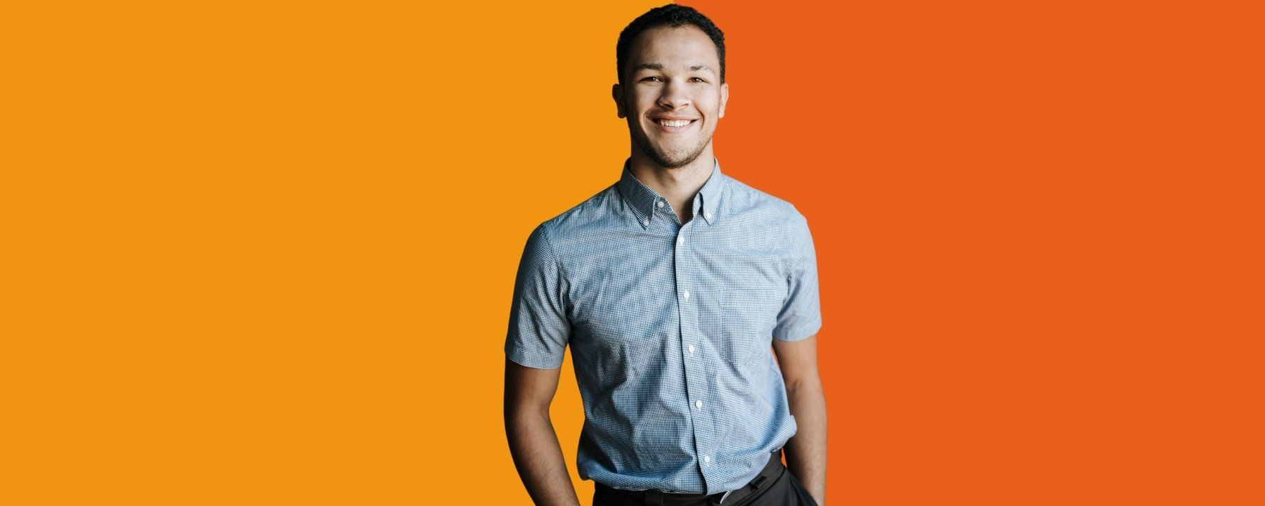 Young man in short sleeved shirt smiling and looking directly at camera. Stands casually with his hands in pocket.