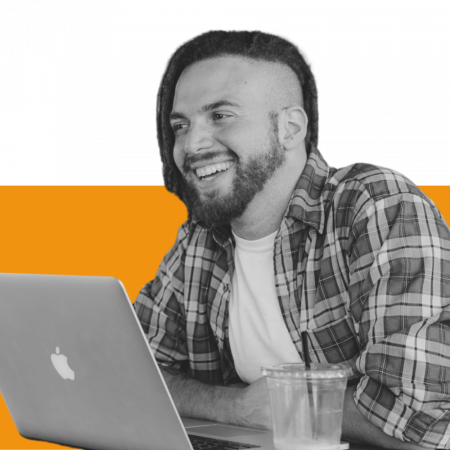 A young man seen in 3/4 profile smiling while sitting in front of a laptop and there is a soft drink container in foreground.