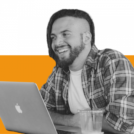 A young man seen in 3/4 profile smiling while sitting in front of a laptop and there is a soft drink container in foreground.